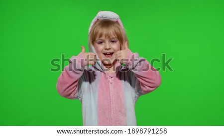 Young little blonde child 5-6 years old smiling, showing thumbs up gesture, agree sign in unicorn costume on chroma key green background. Portrait of kid girl animator in unicorn pajamas
