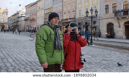 Senior pensioners couple man and woman tourists. Elderly grandfather taking photo pictures with grandmother on retro camera. Family enjoying vacations time together in winter snowy town Lviv, Ukraine