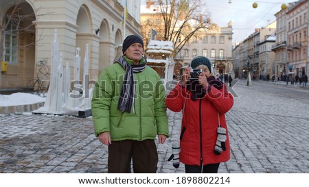 Senior wife and husband tourists taking photo pictures on retro camera, walking, gesturing on winter city street in Lviv, Ukraine. Family vacation activities and photography. Life after retirement