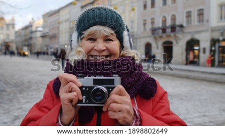 Portrait of old senior woman tourist taking pictures with photo camera, looking at camera using retro device in winter city center of Lviv, Ukraine. Photography, travelling, vacation. Active pensioner
