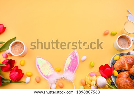 Spring breakfast. Espresso coffee in bright cups, Easter chocolate eggs, pink bunny ears and red tulips on a yellow background. Free space for text. Copy space. Flat lay 