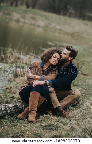 Happy lovers are sitting on the shore of the lake. Young couple is hugging on autumn day outdoors. A bearded man and curly woman in love. Valentine's Day. Concept of love and family