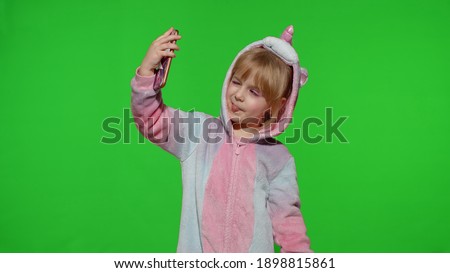 Portrait of blonde kid emotionally makes selfie on mobile phone. Child girl in unicorn pajamas costume using smartphone. Video call, blog, play games, looking at cellphone. Chroma key green background