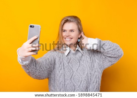 Happy young woman in sweater taking selfie with smartphone and showing thumb up