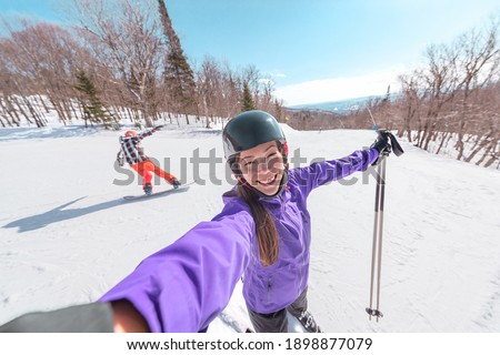 Winter sports fun - Happy Asian skier taking selfie with snowboarder friend in the background. Ski resort slope vacation.