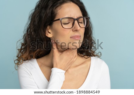 Woman having sore throat, tonsillitis, feeling sick, suffering from painful swallowing, angina, strong pain in throat, loss of voice, holding hand on her neck, isolated on studio blue background. Royalty-Free Stock Photo #1898884549