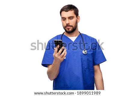 medicine, healthcare and technology concept - doctor or male nurse in blue uniform with stethoscope using smartphone over white background
