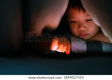 Asian boy hiding in the blanket on bed and playing digital tablet. Soft focus image.