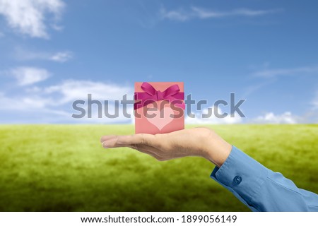 Human hand showing pink gift box with red ribbon and heart with blue sky background. Valentines day