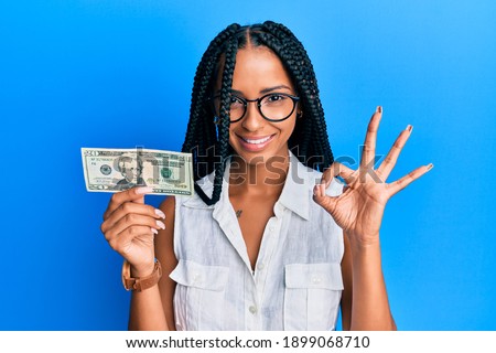 Beautiful hispanic woman holding 20 dollars banknote doing ok sign with fingers, smiling friendly gesturing excellent symbol 