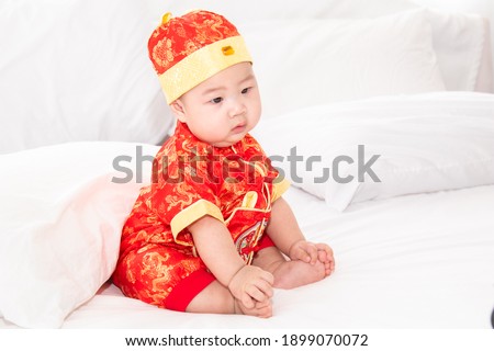 Adorable Asian boy in red Chinese Costume clothes sitting on bed at home looking for somethin with curiosity, innocent cute baby feel sleepy yawning boring waiting for mother feeding