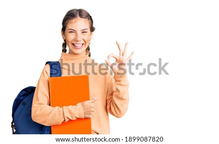 Beautiful caucasian woman with blonde hair wearing student backpack and holding book doing ok sign with fingers, smiling friendly gesturing excellent symbol 