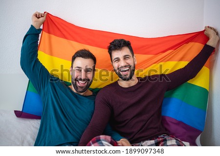 Gay couple holding lgbt rainbow flag indoors on bed at home - Focus on right man face Royalty-Free Stock Photo #1899097348