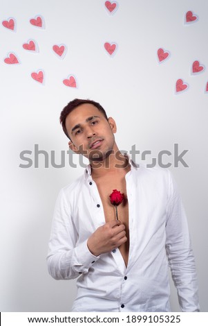Young man looking happy and in love, holding a red rose, with many paper hearts flying behind him, white background. Concept of love, Valentine