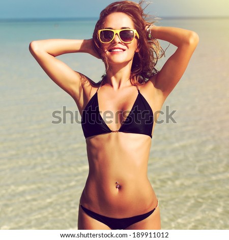 Outdoor summer portrait of pretty young smiling happy woman in sunglasses and black bikini posing near the sea and blue sky