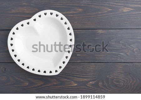 White plate in the shape of a heart on a gray wooden background with space for text. Layout for your postcard .