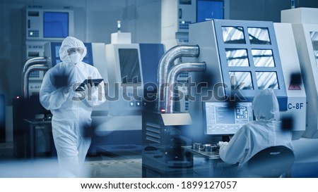 Research Factory Cleanroom: Engineer Wearing Coverall and Mask, Walking Through Workshopop Uses Tablet Computer. In Background Professionals Work on Modern High Tech CNC Machinery Royalty-Free Stock Photo #1899127057
