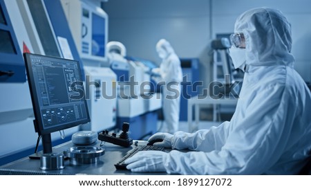 Factory Cleanroom: Engineer Wearing Coverall and Mask Does System Control on a Computer, Uses Controller, Screen Shows Infographics. In Background Modern CNC Machinery, Robot Arm, Electronic Equipment Royalty-Free Stock Photo #1899127072