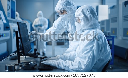 Factory Cleanroom: Engineer and Scientist Wearing Coveralls and Masks Have Discussion, Use Computer Showing Infrastructure System Control. In Background CNC Machinery, Electronics Equipment Royalty-Free Stock Photo #1899127075