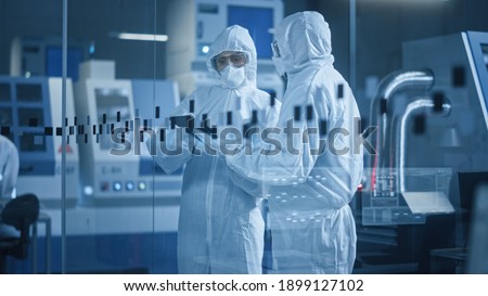 Factory Cleanroom: Engineer and Scientist Wearing Coveralls, Standing in Workshop Talk and Use Tablet Computer, Professionals Develop Technology for Modern CNC Machinery and Electronic Equipment Royalty-Free Stock Photo #1899127102