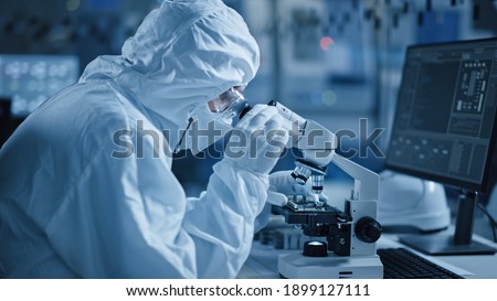 Research Factory Cleanroom: Engineer Scientist wearing Coverall, Gloves Uses Microscope to Inspect Samples, Developing High Tech Modern Technology for Medical and High Precision Electronics Industry Royalty-Free Stock Photo #1899127111