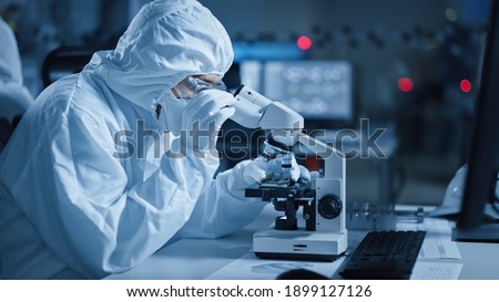 Research Factory Cleanroom: Engineer Scientist wearing Coverall, Gloves Uses Microscope to Inspect Samples, Developing High Tech Modern Technology for Medical and High Precision Electronics Industry Royalty-Free Stock Photo #1899127126