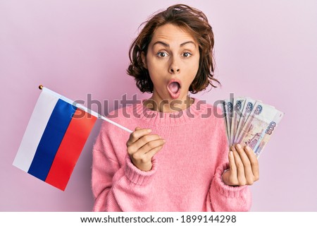 Young brunette woman holding russia flag and rubles banknotes afraid and shocked with surprise and amazed expression, fear and excited face. 