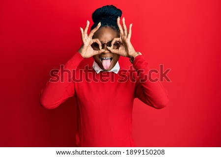 Young african american woman wearing casual clothes and glasses doing ok gesture like binoculars sticking tongue out, eyes looking through fingers. crazy expression. 