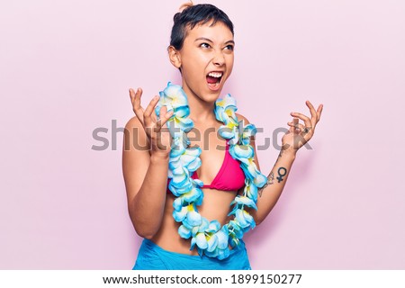 Young woman wearing bikini and hawaiian lei crazy and mad shouting and yelling with aggressive expression and arms raised. frustration concept. 