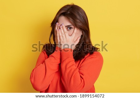 Picture of shy brunette teenage girl covering face with hands and peeping through her fingers at camera. Young attractive female hiding her face being frightened, scared or ashamed, wears casually. Royalty-Free Stock Photo #1899154072