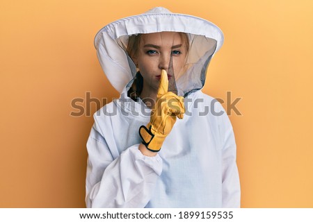 Beautiful blonde caucasian woman wearing protective beekeeper uniform asking to be quiet with finger on lips. silence and secret concept. 