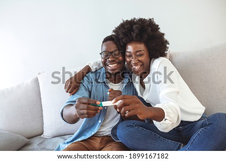 Enamoured couple finding out results of a pregnancy test sitting on the bed. Happy couple checking pregnancy test sitting on a couch in the living room at home. Family, parenting and medical concept