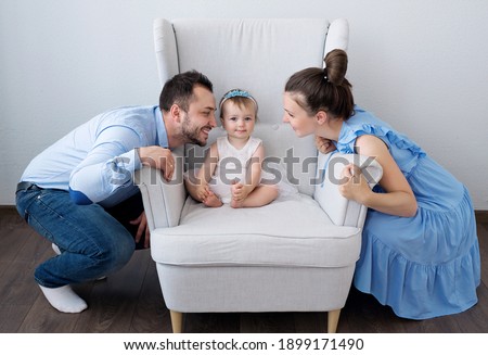 A little girl is sitting on a chair, and her mom and dad want to kiss her. The love of parents is priceless.

