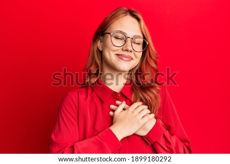 Young beautiful redhead woman wearing casual clothes and glasses over red background smiling with hands on chest with closed eyes and grateful gesture on face. health concept. 