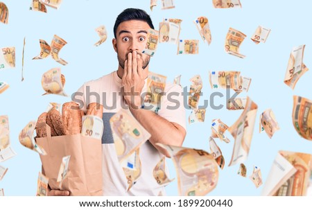 Young handsome man holding bread bag covering mouth with hand, shocked and afraid for mistake. surprised expression
