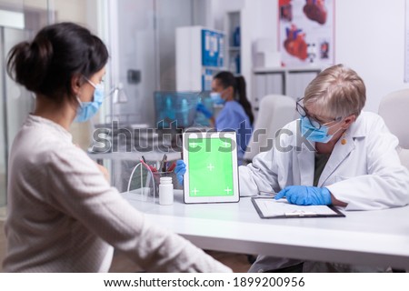 Elderly doctor holding tablet with green screen in clinic room speaking with young patient explaining the treatment looking at mockup display. Showing at izolated monitor, chroma key desktop.