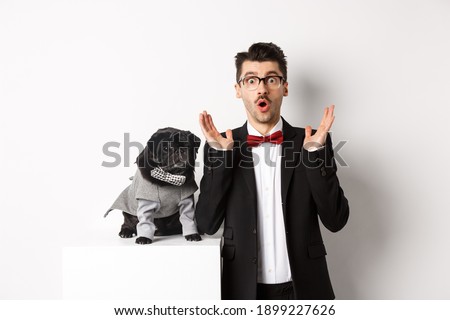 Animals, party and celebration concept. Image of dog owner and cute pug in costumes suits staring surprised at camera, reacting on promo offer, white background