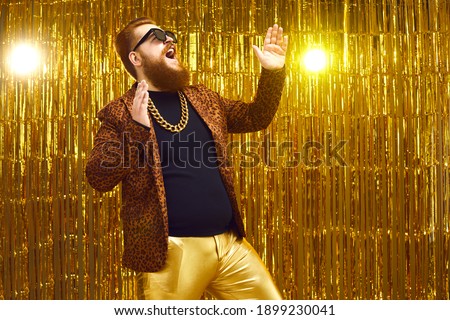Excited rich red-haired young man in neck chain and cool glasses having fun at glamour night club disco party. Happy fashionable bearded showman singing and dancing on stage with golden background