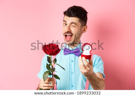 Valentines day. Funny guy making proposal, winking and saying marry me, showing engagement ring with red rose, standing over pink background Royalty-Free Stock Photo #1899233032