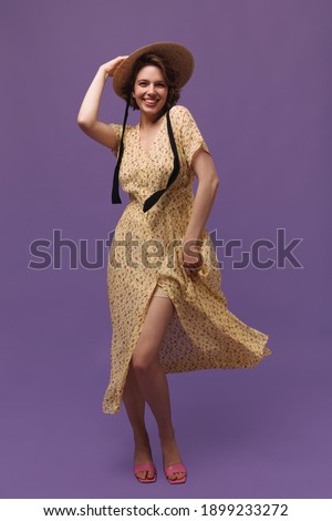 Portrait of young lady in yellow dress, posing against violet background. Handsome dark-haired girl with floral outfit, straw hat and pink shoes looking into camera, smiling and holding skirt