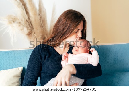 Young lovely mom holds a newborn baby and kisses the baby tenderly. A woman and a child are sitting on a blue sofa, in the background beige shades and an reed.