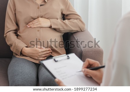 Young asian pregnant woman holding her belly while gynecologist notes the symptoms that the pregnancy is explaining about the unborn child. Royalty-Free Stock Photo #1899277234