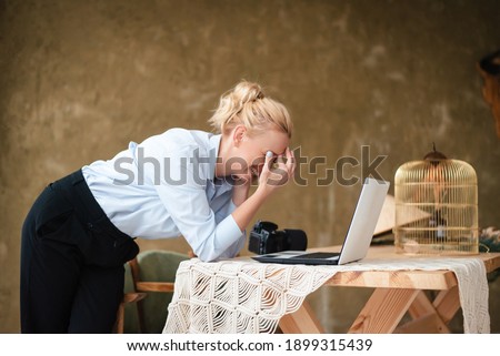 Smiling young woman leaning with elbows on wooden table and looking at laptop screen. Female photographer enjoying favorite work at home.