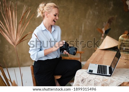 Side view of cheerful young woman with blond hair holding digital camera and looking at computer screen while working at home. Creative lifestyle.