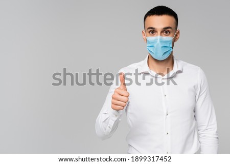 brunette hispanic businessman in medical mask showing thumb up isolated on grey