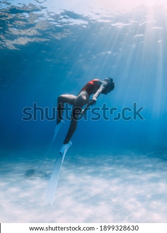 Woman freediver in red swimsuit with white fins glides underwater over sand in tropical ocean. Sporty girl dive underwater.