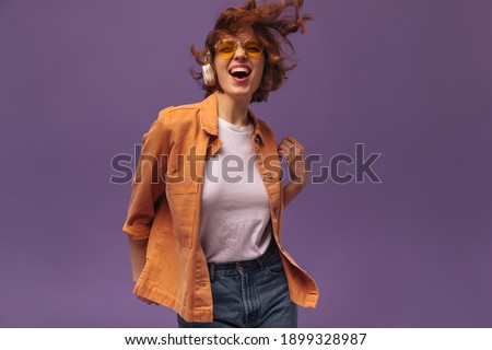 Expressive young brunette dancing against violet background. Attractive dark-haired curly girl in orange shirt, white top and stylish glasses, wearing headphones, listening music and enjoying