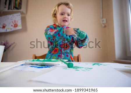 Cute blonde girl paitining with many colors using paintbrush and fingers.