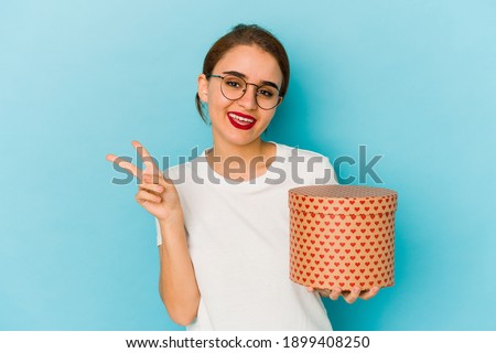 Young skinny arab girl holding a valentines day box joyful and carefree showing a peace symbol with fingers.