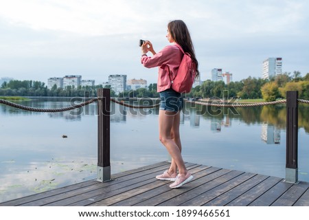 girl in summer in a city park near reservoir, pond, river and lake, records video photos on smartphone. Sightseeing and travel. Landscape photography. Denim shorts jacket backpack